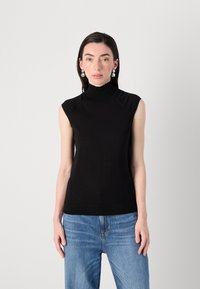 Modelka má na sobě bezrukávový černý rolák a světle modré džíny, pózuje na jednolitém bílém pozadí. Elegantní, minimalistický styl.