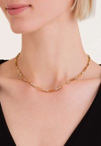 Ogrlica od metalne legure u zlatnoj boji koja sadrži izdužene karike i malo, prozirne ukrasne kamenčiće ravnomjerno raspoređene. Dizajnirana da leži na ključnoj kosti.