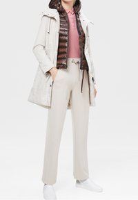 Veste d'extérieur beige clair, avec capuche et cordons de serrage. Veste matelassée intérieure marron, polo rose, et pantalon beige clair avec ceinture. Chaussures blanches.