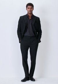 Schwarzer, maßgeschneiderter Anzug mit einem doppelt geknöpften Sakko, kombiniert mit einem dunkel gemusterten Hemd und schmal geschnittenen Hosen. Schwarze Lederstiefel vervollständigen den Look.