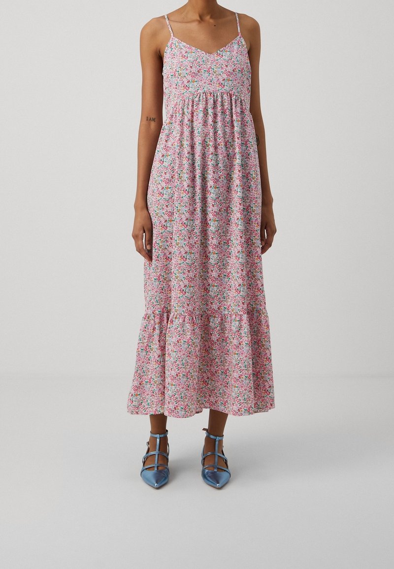 Žena ve růžových květinových midi šatech bez rukávů s tenkými ramínky a modrými lodičkami s ankletovým páskem na jednobarevném pozadí.