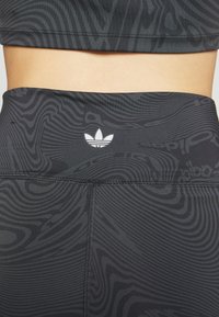Černý sportovní crop top a legíny s vysokým pasem, které mají vířivé texturované vzory, s bílým logem Adidas umístěným uprostřed na pasovém lemu.
