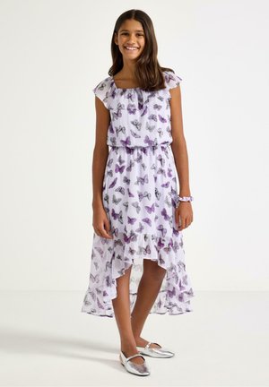 Hymyilevä tyttö, joka pukeutunut hihattomaan valkoiseen mekkoon, jossa on violetteja perhoskuvioita, ja hopeisiin litteisiin kenkiin, seisoo yksinkertaista valkoista taustaa vasten.