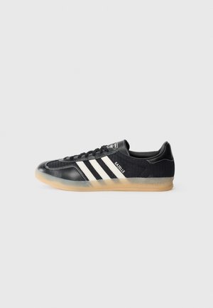 Czarny sneaker Adidas Gazelle z białymi paskami, gumową podeszwą i sznurowaniem, pokazany z boku na szarym tle.