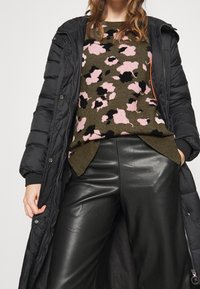 Donna che indossa un lungo cappotto trapuntato nero, un maglione verde oliva con motivo floreale astratto rosa e nero, e pantaloni in pelle neri.