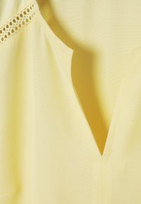 Tissu jaune clair avec encolure en V et bordure en dentelle décorative le long de la couture de l'épaule, montrant une texture lisse et des détails de couture.