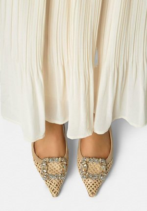 Voeten met beige geweven puntige flats met grote rechthoekige strassgespen onder een lange witte geplooide rok.