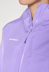 Fioletowa lekka kurtka z wysokim kołnierzem, wyposażona w zamek błyskawiczny z przodu oraz logo "MAMMUT" w białym kolorze na obszarze klatki piersiowej. Gładka tekstura.