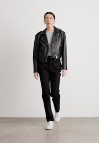 Giacca nera in pelle cropped con zip argentate, maglietta grigia, pantaloni neri e sneakers bianche. Modella che cammina su uno sfondo neutro.