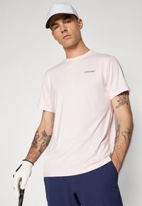 Svijetlo ružičasta majica kratkih rukava s Calvin Klein logotipom na lijevoj strani grudi, u kombinaciji s tamnoplavim hlačama i bijelom kapom. Vidljive tetovaže na ruci.