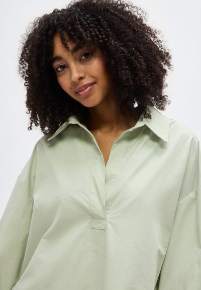 Νεαρή γυναίκα με σγουρά μαλλιά φοράει ένα άνετο, ανοιχτοπράσινο πουκάμισο με γιακά, χαμογελώντας απαλά μπροστά σε ένα απλό φόντο.