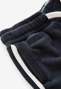 Pantaloni da tuta blu navy con una texture morbida, vita elastica, strisce laterali bianche e un sottile dettaglio sulla tasca.