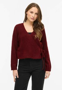 Bordo spalvos megztinis su užrišamu liemeniu, pagamintas iš minkštos, tekstūruotos medžiagos, su ilgomis rankovėmis. Kirptas ties juosmeniu, turintis gilią V formos iškirptę.