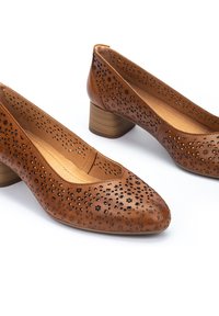 Escarpins en cuir marron avec un design floral perforé, bout pointu et un talon en bois de 2,5 cm. Texture lisse et accents minimalistes.