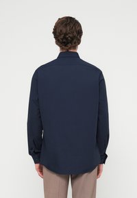 Άνδρας με σγουρά μαλλιά που φοράει μακρυμάνικο σκούρο μπλε πουκάμισο και ανοιχτό καφέ παντελόνι, γυρισμένος πλάτη σε ένα απλό λευκό φόντο.