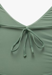 Maillot de bain vert avec une texture douce et lisse. Présente un décolleté en V profond et des liens réglables à l'arrière, avec des détails cousus le long des bords.
