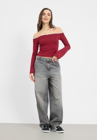 Červený top bez ramienok s dlhými rukávmi, skombinovaný s šedými širokými džínami. Model nosí čierne tenisky s bielymi detailmi.