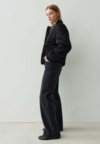 Femme portant une veste en quilt noir, un jean noir à jambes larges et des chaussures noires, debout de côté les mains dans les poches contre un fond uni.