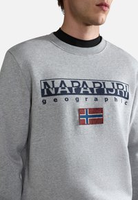 Il pullover grigio presenta un logo blu con la scritta "NAPAPIJRI geographic" e una patch ricamata con la bandiera norvegese. Design a girocollo, tessuto misto cotone.