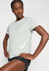 Svetlozelené športové tričko s krátkymi rukávmi, na boku s bielym logom Nike swoosh. Hladká textura a jemný lesk. Čierne šortky.