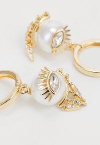 Boucles d'oreilles en or en forme de créoles, ornées d'une grande perle et d'un motif œil décoratif avec des accents de cristal et des détails en piques. Surface lisse et finition brillante.