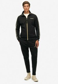 Chándal negro con cremallera y detalles dorados. Presenta mangas largas, rayas laterales y pantalones ajustados. Combinado con zapatillas altas beige.