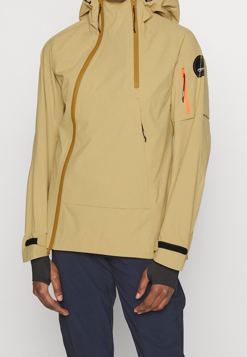 Veste à capuche beige avec fermeture éclair asymétrique à l'avant, fermeture éclair orange sur la poche de manche, sangles noires aux poignets, portée avec un pantalon sombre sur une personne.