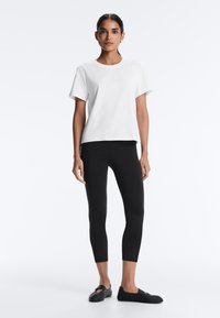 Woman standing, wearing a plain white short-sleeve shirt, black cropped leggings, and black slip-on shoes against a white background.