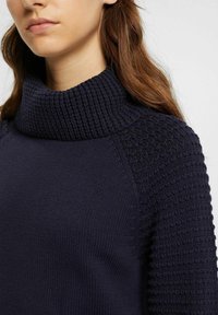 Pull en maille bleu marine avec un col haut texturé et un motif nid d'abeille sur les épaules, présentant un corps lisse et des accents côtelés.