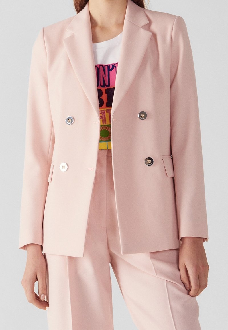 Femme portant un blazer rose clair croisé et un pantalon assorti avec un t-shirt graphique coloré en dessous.