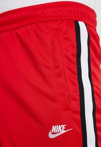 Red athletic shorts made of smooth fabric with a side pocket, featuring black and white vertical stripes and a white Nike logo.
