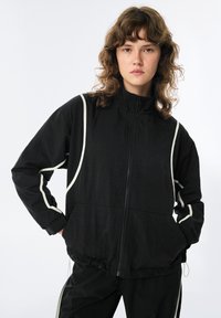 Giacca nera leggera con colletto alto, caratterizzata da piping bianco lungo le cuciture, un fondo elastico e chiusura frontale con zip.