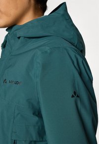 Jachetă impermeabilă de culoare teal, cu guler înalt, glugă reglabilă și accente negre cu logo pe umăr și piept. Textură netedă și ușoară.