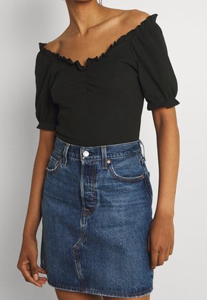 Personne portant un haut noir à épaules dénudées avec manches bouffantes et une jupe en jean bleue taille haute sur fond uni.