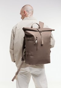 Brun rull-up ryggsäck gjord av hållbart material. Utrustad med justerbara remmar, en sidoficka med dragkedja och en texturerad yta. Minimalistisk design.