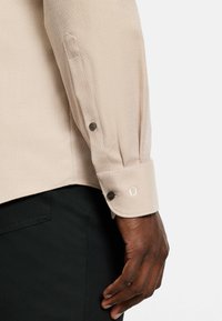 Eine dunkelhäutige Hand ruht neben einer schwarzen Hose und trägt ein beige-langärmeliges Hemd mit kleinem Diamantmuster und dunklen Knöpfen an der Manschette und im Ärmel.