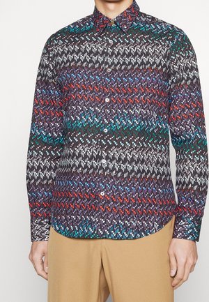 Man draagt een shirt met lange mouwen en knopen, met een kleurrijk abstract patroon en beige broek, staand voor een effen achtergrond.