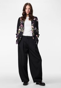 Woman with long wavy hair wearing a white shirt, floral black blazer, wide black pants, and black shoes, standing against a plain background.