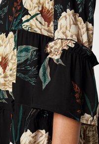 Svart blommig klänning med stora beige blommor, gröna blad och orange detaljer. Ärmarna har rynkade detaljer och elastiska fållar.