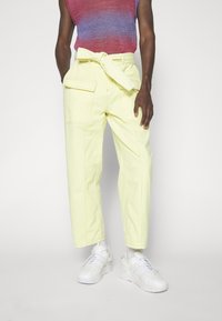 Persona que lleva pantalones de tiro alto de color amarillo pálido con cinturón de lazo y grandes bolsillos, zapatillas blancas y una blusa sin mangas de color rojo-violeta.
