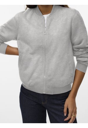 Person wearing light gray zip-up bomber jacket with ribbed cuffs and dark blue jeans, posing with one hand on hip and the other relaxed.