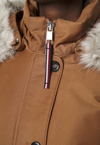 Giacca bomber marrone con cappuccio foderato in pelliccia, dotata di cerniera argentata, chiusura con bottoni e un accento a strisce sul tiralampo. Tessuto morbido.