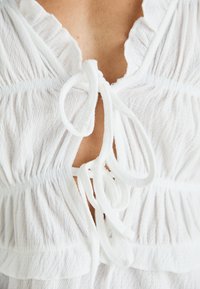 White textured top featuring a ruffled neckline, adjustable tied detail at the front, and gathered fabric for a relaxed fit.