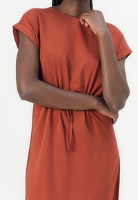 Šaty v rezavé barvě z lehkého materiálu. Krátké ohrnovací rukávy a nabíraný pas s detailem na zavázání. Hladká textura a jednoduchý design.