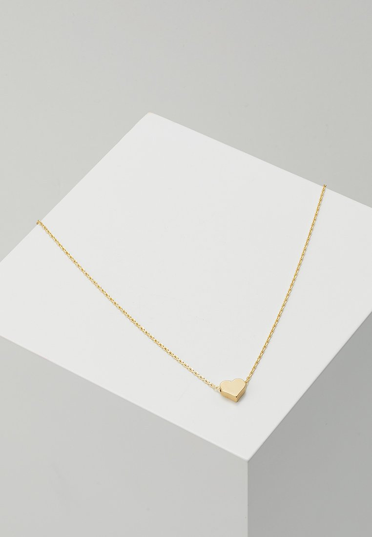 Collier en chaîne de couleur dorée avec un pendentif en forme de cœur à la surface lisse. Le design est délicat et minimaliste.