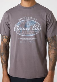 Grå bomulds t-shirt med et globusmotiv og teksten "Closure Ldn" i hvid og blå, korte ærmer og rund halsudskæring.