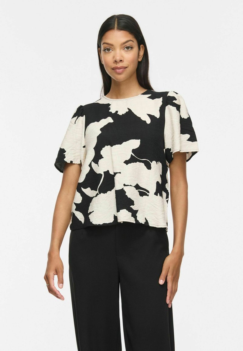 Femme portant un chemisier à manches courtes noir et blanc à motifs avec des manches bouffantes et un pantalon noir, debout devant un fond blanc uni.