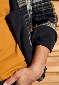 Giacca in pile nera con rivestimento a quadri, indossata sopra una t-shirt color senape. Mano visibile poggiata in tasca, sfondo in legno.