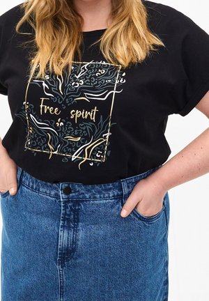 Kobieta nosząca czarny t-shirt z tekstem "Free spirit" i abstrakcyjnym wzorem, włożony do wysokiej talii niebieskiej dżinsowej spódnicy, dłonie w kieszeniach.