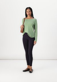 Grønn langermet topp med avslappet passform, kombinert med svarte tights. Modellen holder en liten brun håndveske. Gulv mot hvit bakgrunn.
